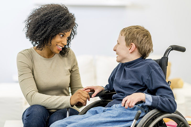 Middle age African American woman is smiling and kneeling next to a smiling white male boy sitting in a wheelchair