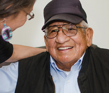 Native American senior man with a baseball cap is smiling while a woman leans over talking to him