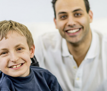 Young boy sitting in a wheelchair and smiling while a 20 to 30 year old man kneels behind him and is smiling
